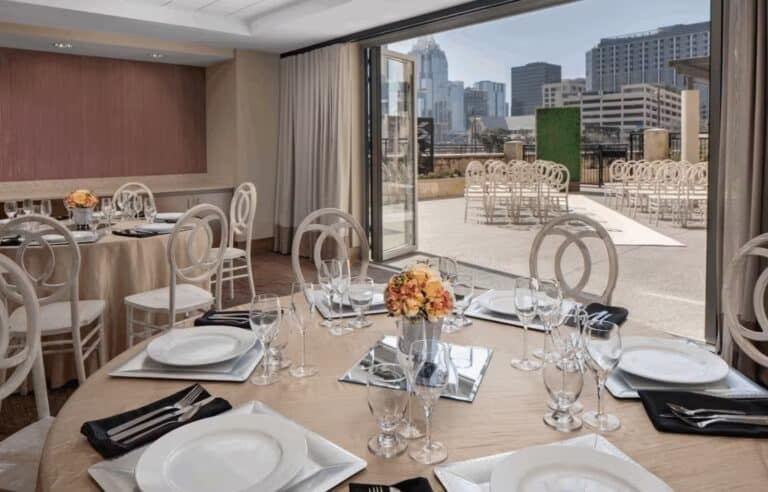 Elegant indoor banquet setup with round tables, white chairs, and floral centerpieces, opens to an outdoor ceremony area with rows of chairs and a city skyline view in the background.